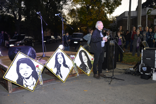 Maestro de ceremonias hablando junto al escenario con las placas de las tres jóvenes asesinadas.